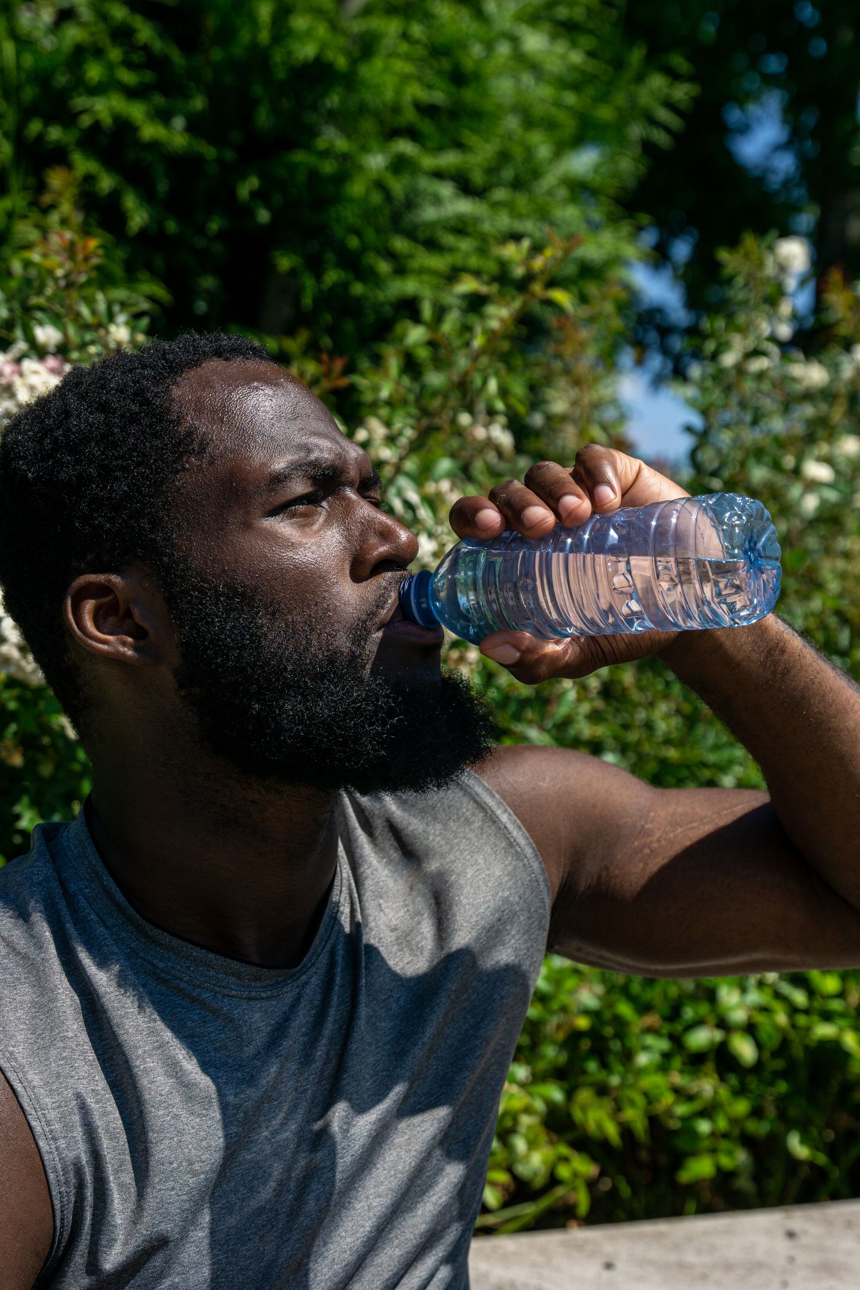 Athletic man drinking water from plastic bottle in sunny outdoor setting.